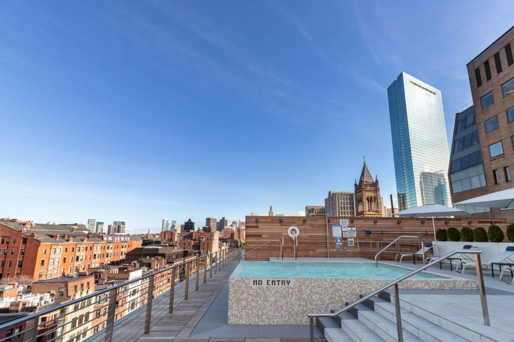 Rooftop pool with lounge chairs, umbrellas, and city skyline in the background on a sunny day at 28 Exeter at Newbury in Boston, Massachusetts