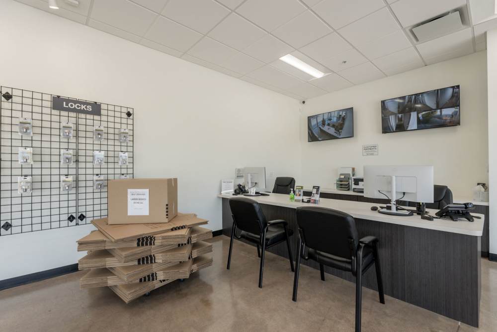 Office reception area with a counter, chairs, security monitors, and a wall display of locks next to stacked cardboard boxes