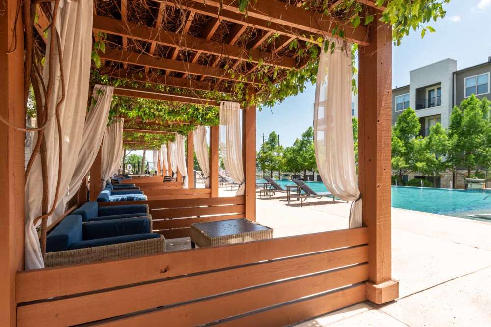 Outdoor pool area with wooden cabanas, blue cushions, and nearby apartment buildings at Henley at The Rim in San Antonio, Texas