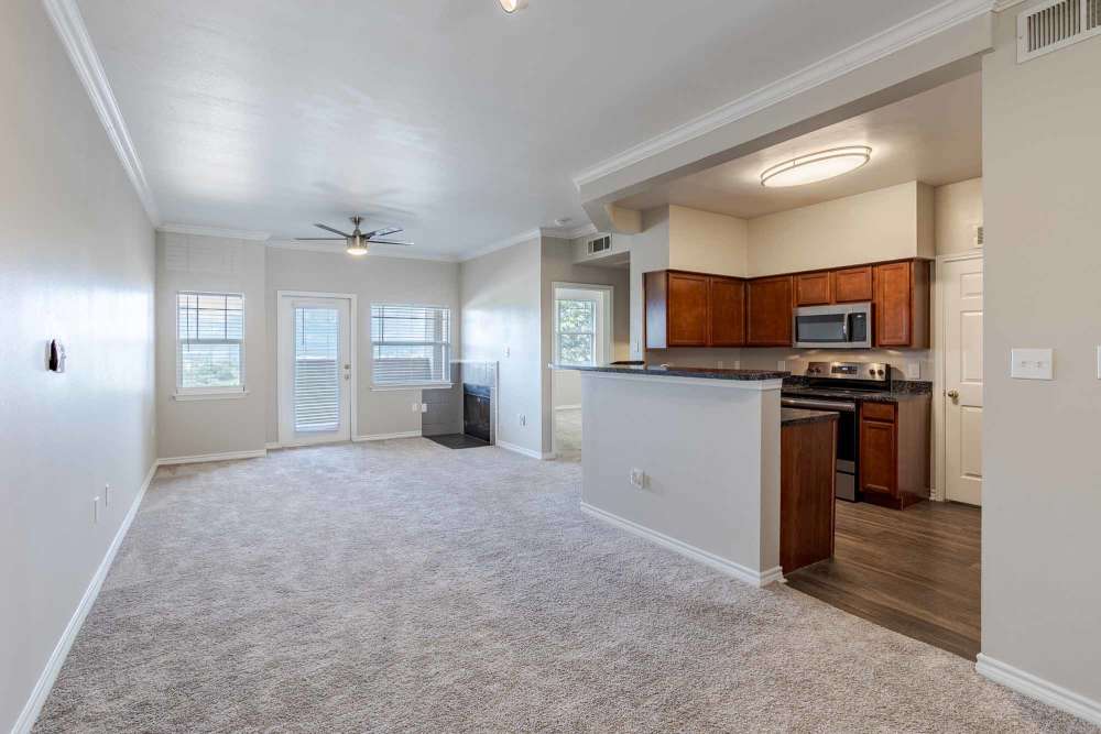 Spacious living area with plush carpeting, bright windows, and an open kitchen design featuring modern appliances, at Bella Springs Apartments in Colorado Springs, Colorado.