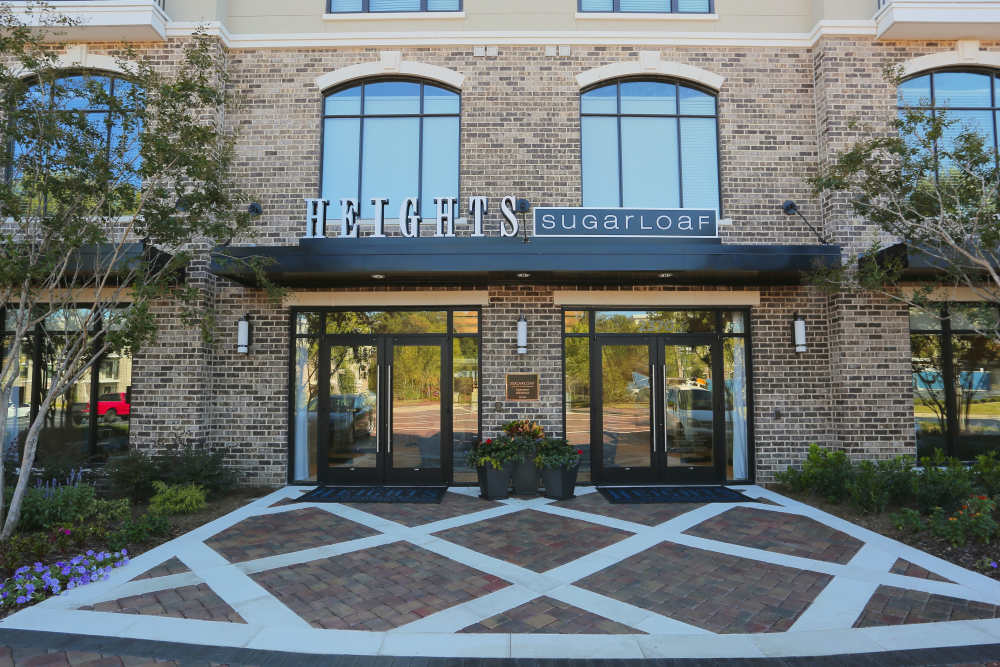 Charming entrance to Heights Sugarloaf, featuring elegant brickwork, vibrant landscaping, and inviting glass doors that welcome you home at 2370 Main at Sugarloaf in Duluth, Georgia
