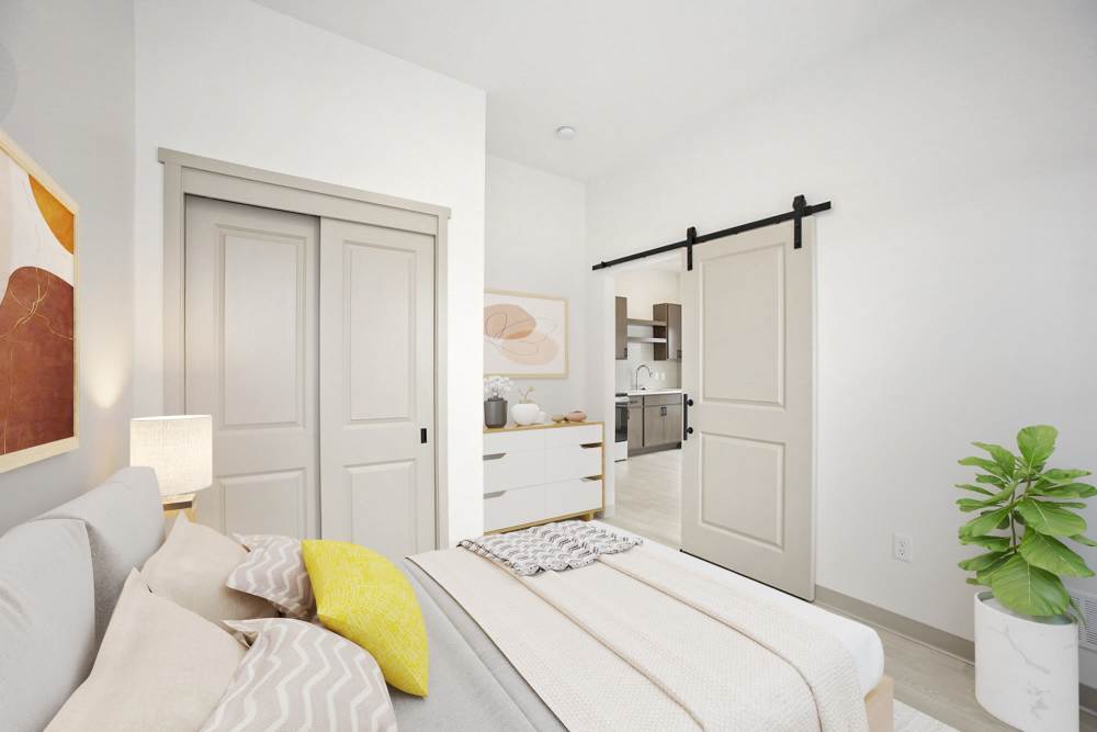 Cozy bedroom with neutral bedding, sliding closet doors, sliding door to kitchen, and potted plant at Terra Station, Hudsonville, Michigan