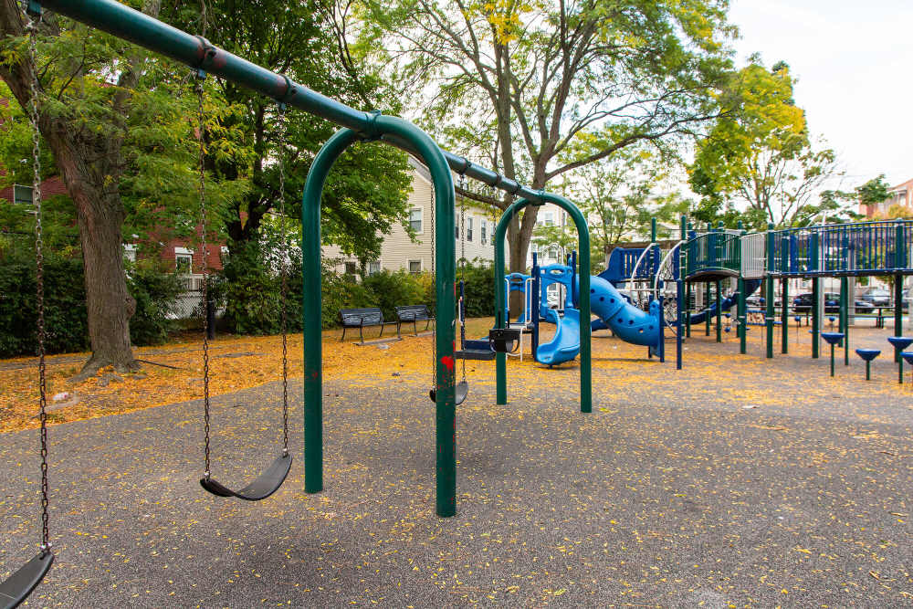 Playground with swings, slide, climbing structures, and autumn leaves, surrounded by trees and buildings at The Lighthouses in Salem, Massachusetts