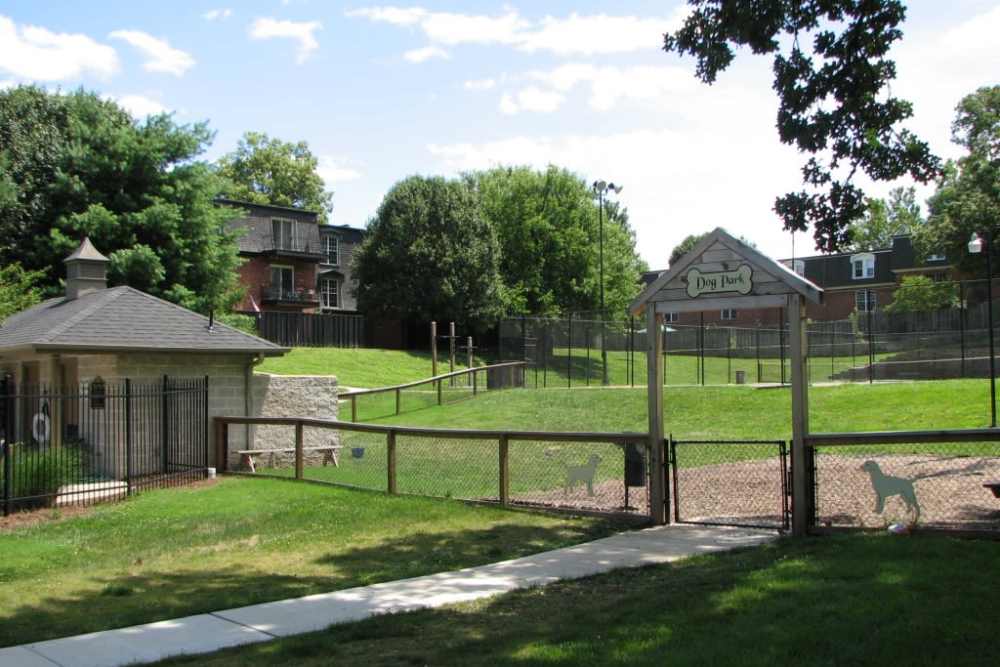 Dog park entrance with wooden archway, decorative dog silhouettes on the gate, grassy area, and trees with homes at Beau Jardin Apartments in St. Louis, Missouri 