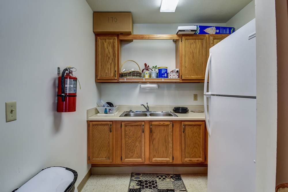 Compact clubhouse kitchen with wooden cabinets, white refrigerator, and sink area at Muirwood Greene in Romney, West Virginia