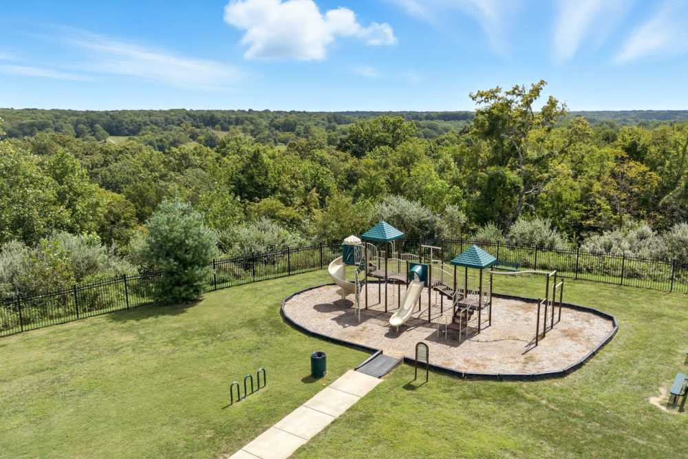 Playground with slides and swings surrounded by green grass and trees, fenced for safety at Bristol Bluffs Apartments in Louisville, Kentucky