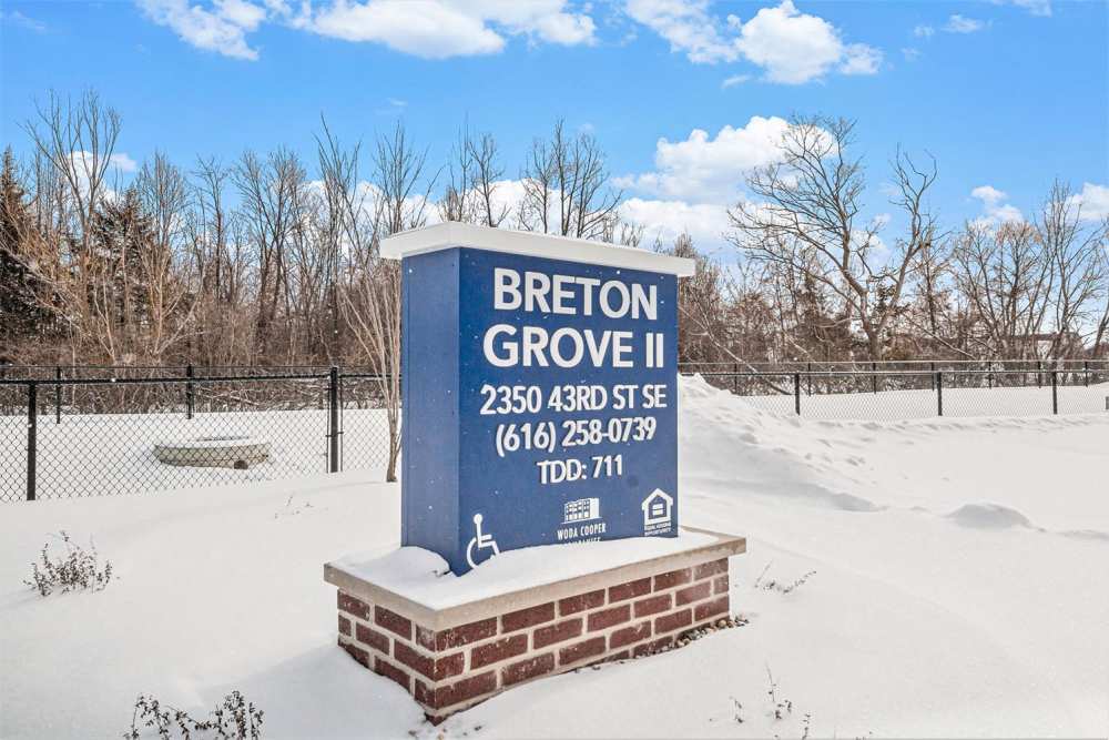 Snow-covered entrance sign for Breton Grove II with trees in the background and a fenced area at Breton Grove II in Grand Rapids, Michigan