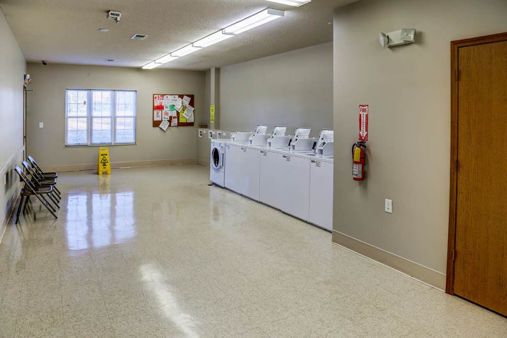 Bright laundry room with multiple washers, chairs, bulletin board, and caution sign on polished floor at Mason Greene in Monticello, Kentucky