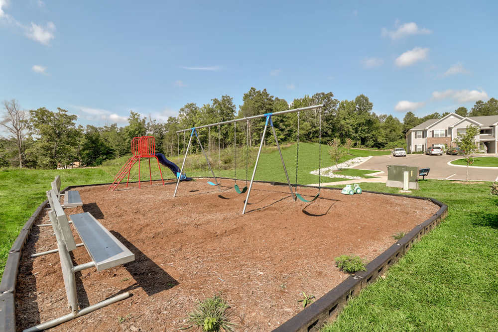 Playground area with swings and slide, benches nearby, green grass and trees in background at Oliver Crossing in Linden, Tennessee