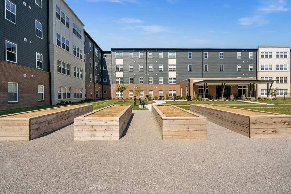 Outdoor community garden beds with modern apartment buildings in the background and seating areas visible at Sparrow Ridge in Hebron, Kentucky