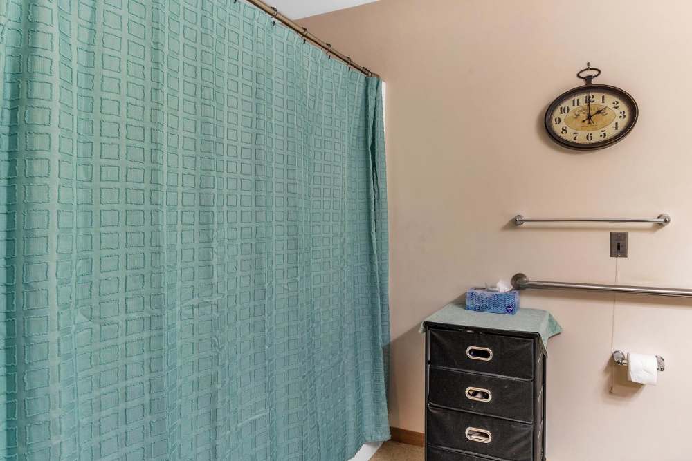 Bathroom with teal shower curtain, wall clock, and storage cabinet with towels and tissue at Tibbits Greene Senior Living in Coldwater, Michigan