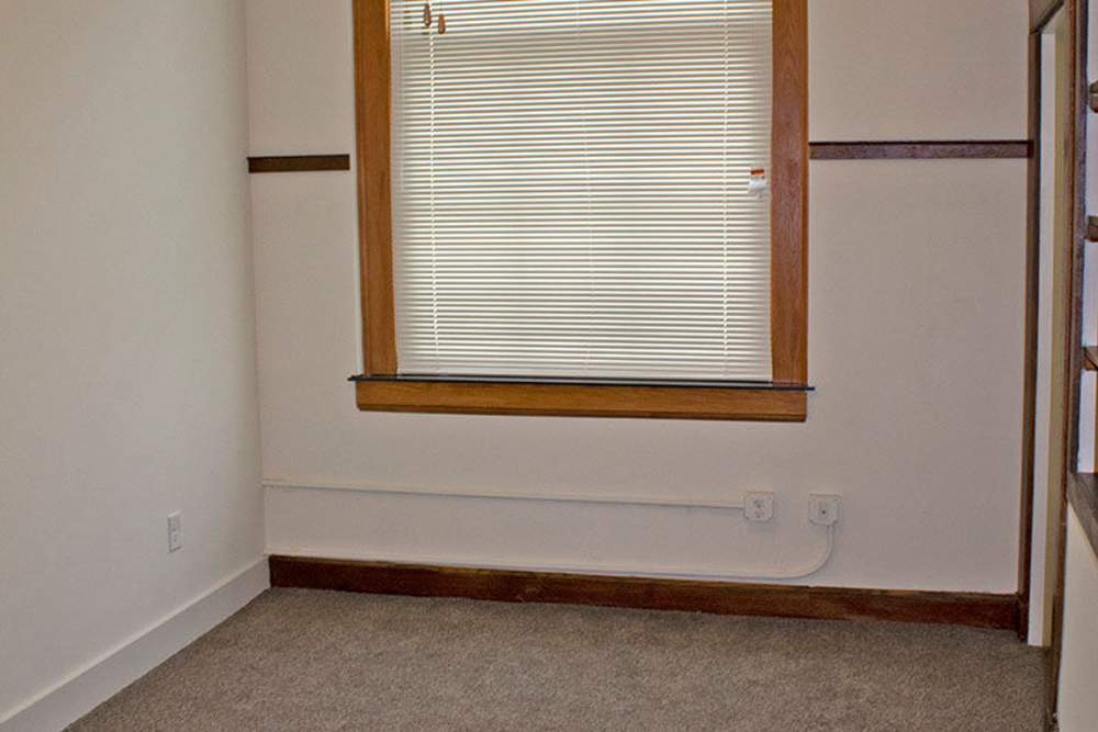 Empty room with beige carpet, wooden window trim, and blinds, featuring a single window at Lincoln House in Owosso, Michigan