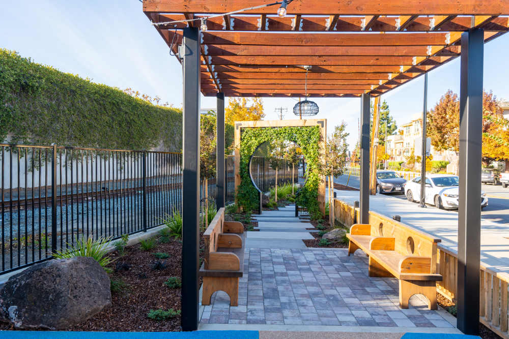 Outdoor seating area with wooden benches and landscaped greenery under a wooden pergola at Exhibit at J Town in San Jose, California