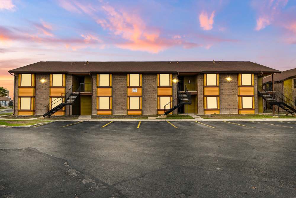 Two-story apartment building with yellow accents and parking lot under a colorful sunset sky at Towne East Village Apartment Homes in Converse, Texas
