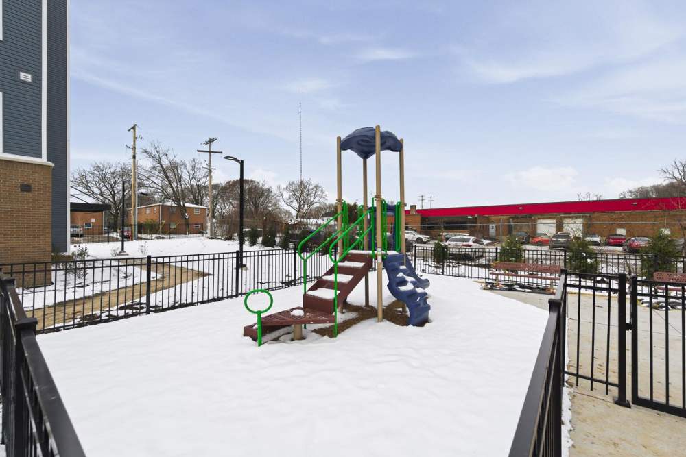 Snow-covered playground with green and brown equipment and fenced area at Granville Woods Lofts in Columbus, Ohio