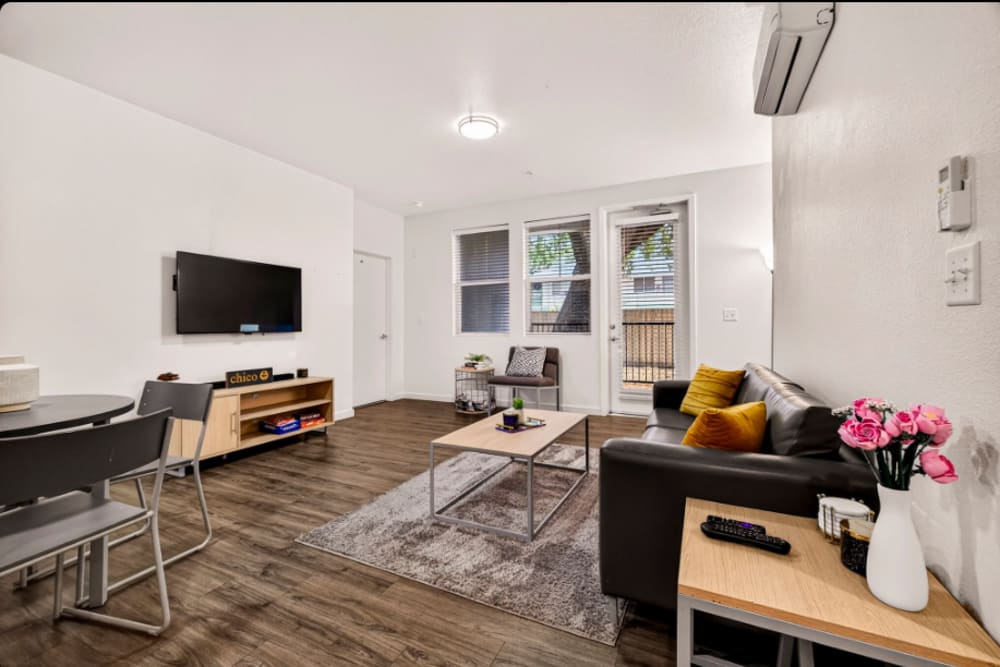 Living room: Bright living room with sofa, wall-mounted TV, coffee table, dining nook, and glass door to balcony at Cedar Flats in Chico, California  