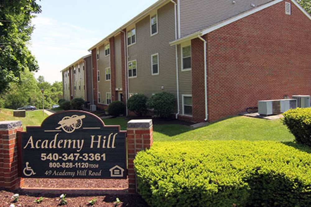 Brick apartment building with landscaped grounds and a welcoming sign in front at Academy Hill in Warrenton, Virginia