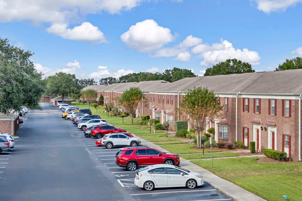 Parking lot with various cars, landscaped lawns, and brick apartment buildings under a blue sky at Carlton Arms Of Winter Haven in Winter Haven, Florida
