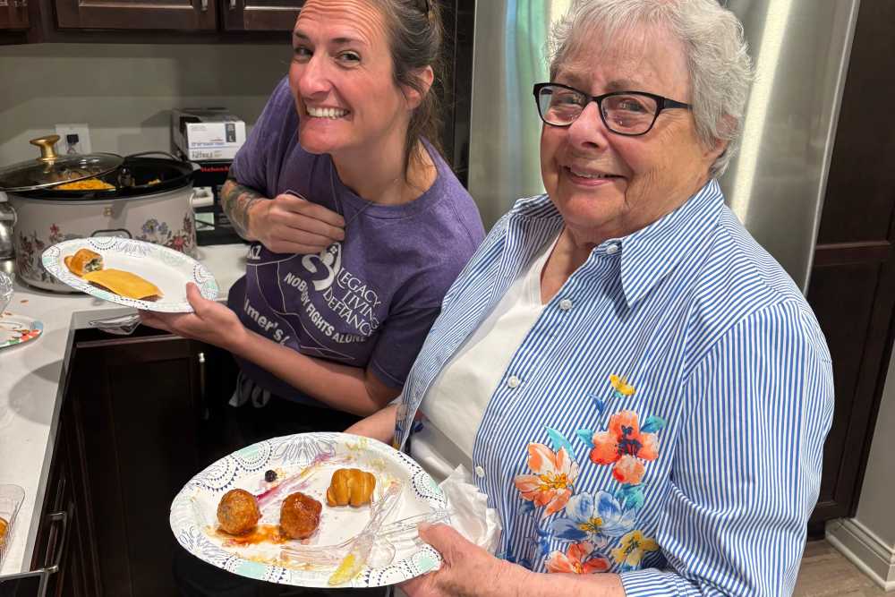 Two people in a kitchen holding plates of food, with a counter of prepared dishes and a slow cooker in the background at Legacy Living Defiance in Defiance, Ohio  