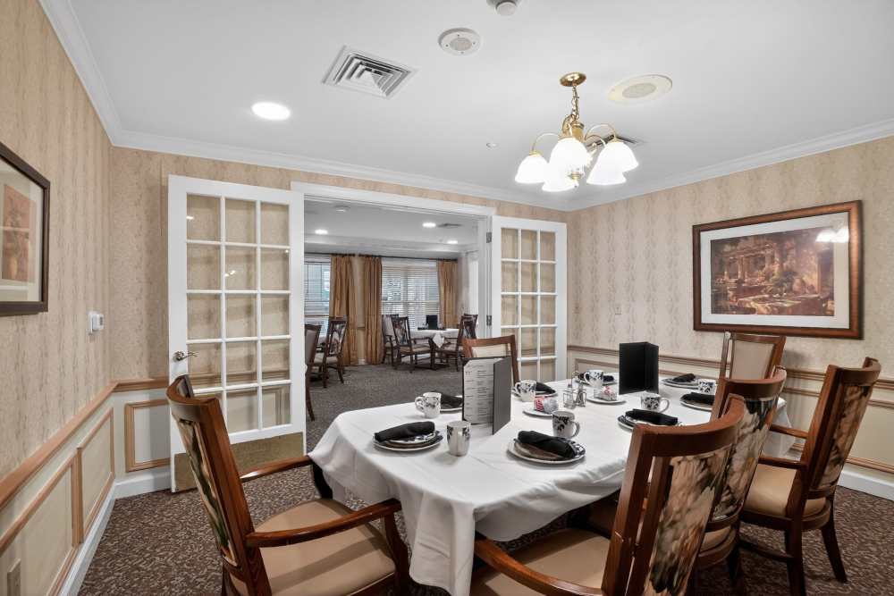 Private dining room with a round table set with dishes and mugs, upholstered chairs, and glass-paneled double doors opening to a larger dining space at Glenncrest at Decatur in Decatur, Georgia