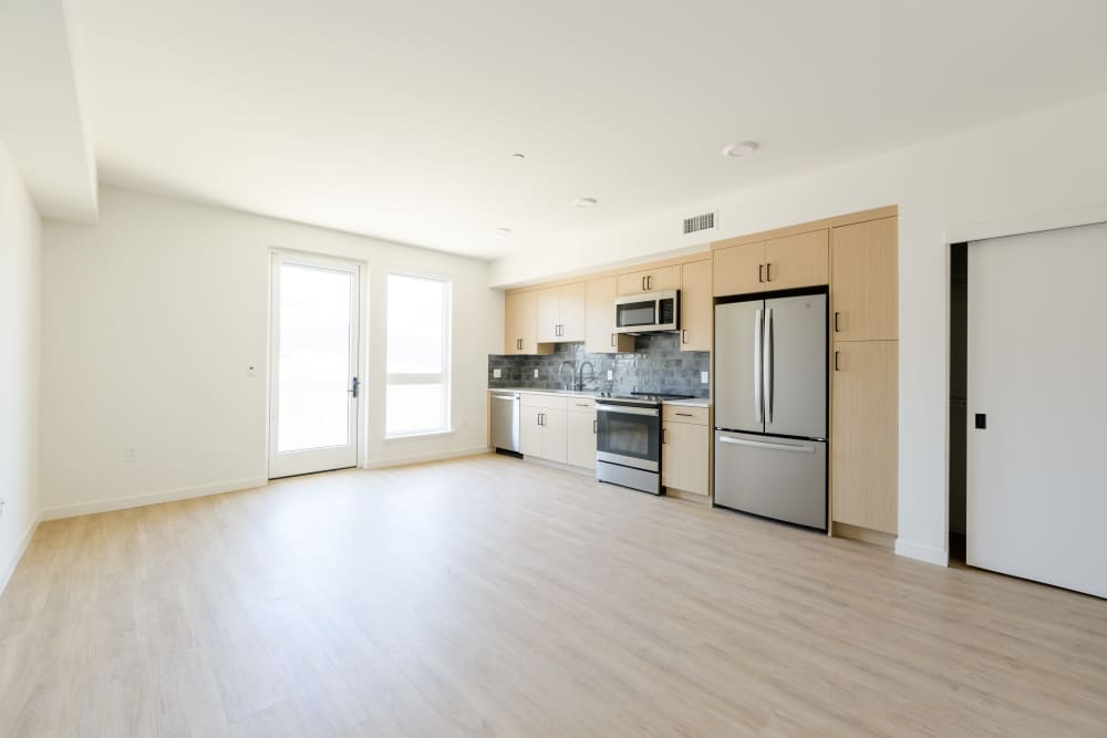Open living area with light wood floors, large window, glass door, and modern kitchen with stainless appliances and wood cabinets at Artisan Crossing in Belmont, California