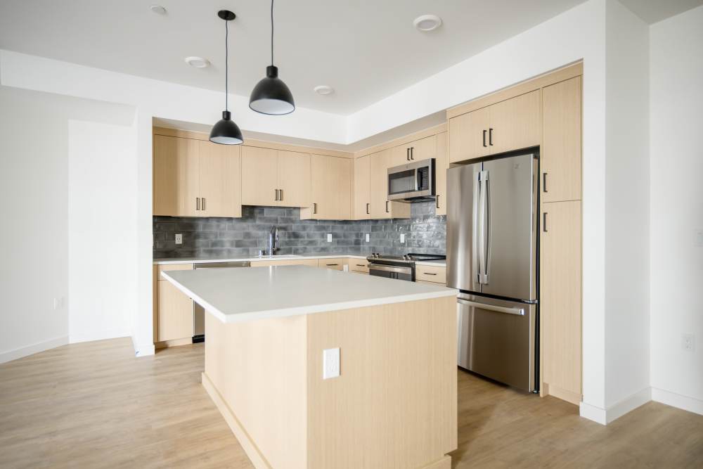 Open kitchen with island, light wood cabinetry, stainless fridge, stove and microwave, gray backsplash, two pendant lights at Artisan Crossing in Belmont, California