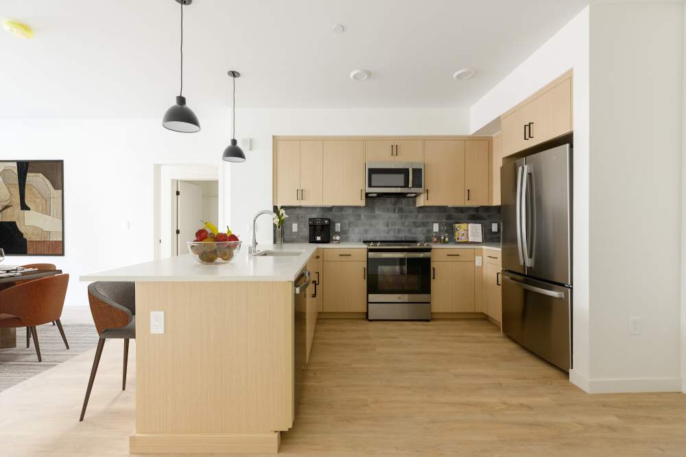 Modern kitchen with island, fruit bowl, pendant lights, wood cabinets, stainless appliances, and gray tile backsplash at Artisan Crossing in Belmont, California