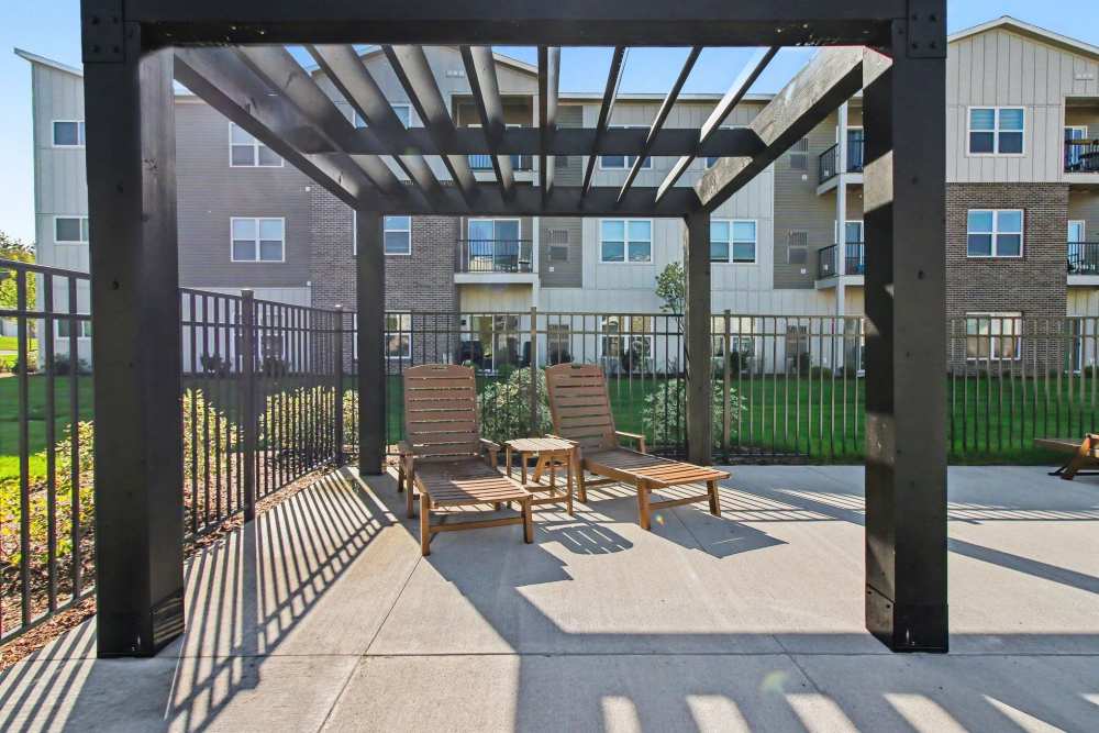 Wooden pergola structure over a concrete patio with two lounge chairs and a small side table, overlooking a grassy area and apartment buildings at The Block at Petoskey in Petoskey, Michigan