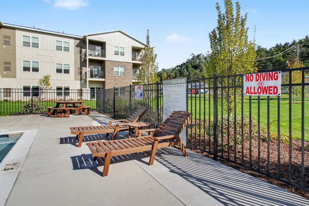 Poolside area with brown chaise lounge chairs, a picnic table, and pool rules signage posted on the black metal fence, with 