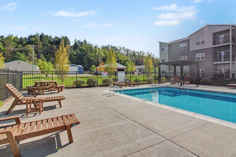 Rectangular swimming pool with chaise lounge chairs and a picnic table on the deck, bordered by a black metal fence, trees, and outbuildings in the distance at The Block at Petoskey in Petoskey, Michigan