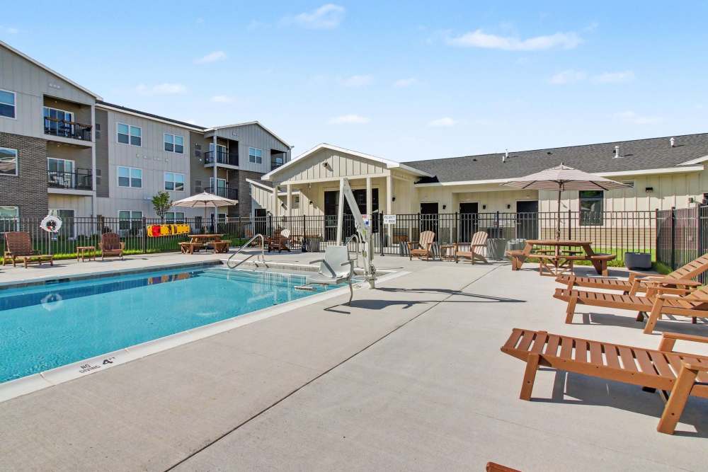 Pool area with a pool lift chair for accessibility, chaise lounge chairs, picnic tables, and striped umbrellas, framed by apartment buildings and a clubhouse at The Block at Petoskey in Petoskey, Michigan