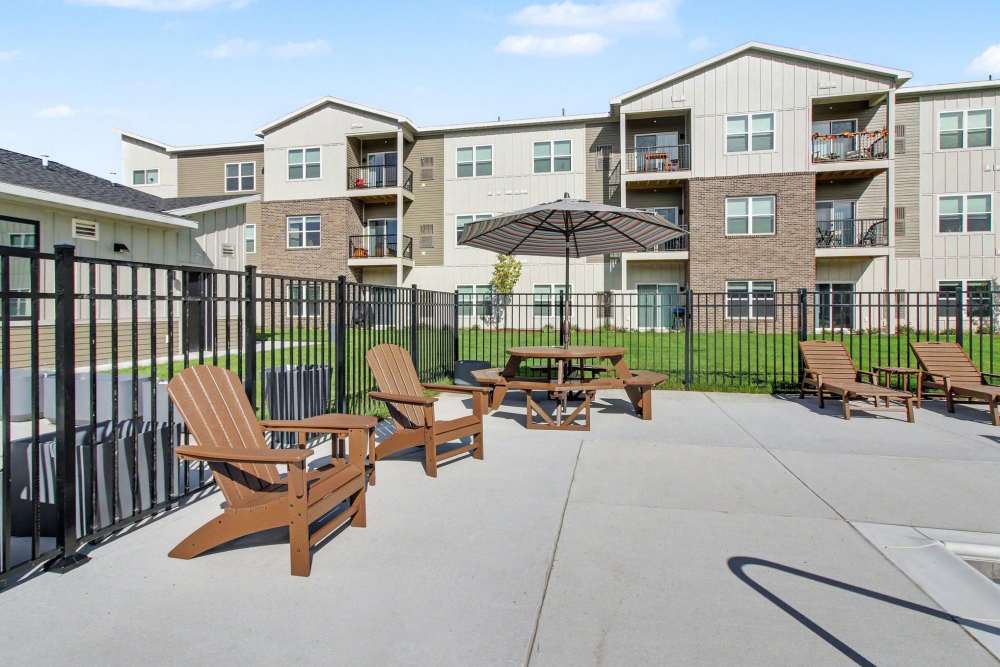 Patio area with two Adirondack chairs, a picnic table with umbrella, and lounge chairs, enclosed by a black fence with apartment buildings and green lawn in the background at The Block at Petoskey in Petoskey, Michigan