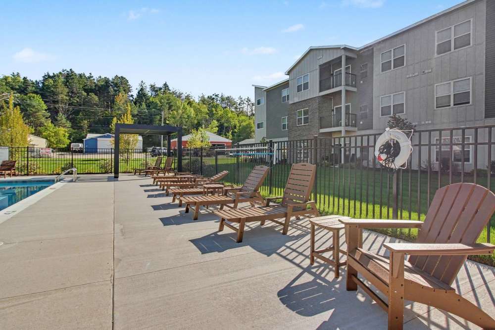 Row of brown chaise lounge chairs beside a pool, with a black pergola frame, safety life ring on the fence, and apartment buildings in the background at The Block at Petoskey in Petoskey, Michigan