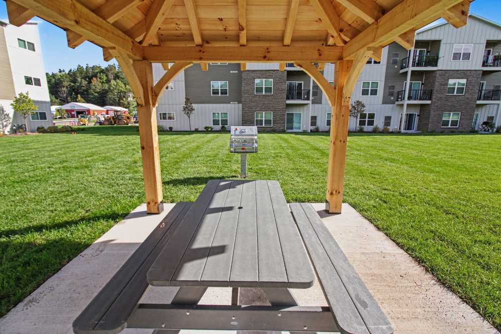 View from inside a wooden gazebo looking out at a gray picnic table, a grill station, and an open lawn with apartment buildings in the background at The Block at Petoskey in Petoskey, Michigan