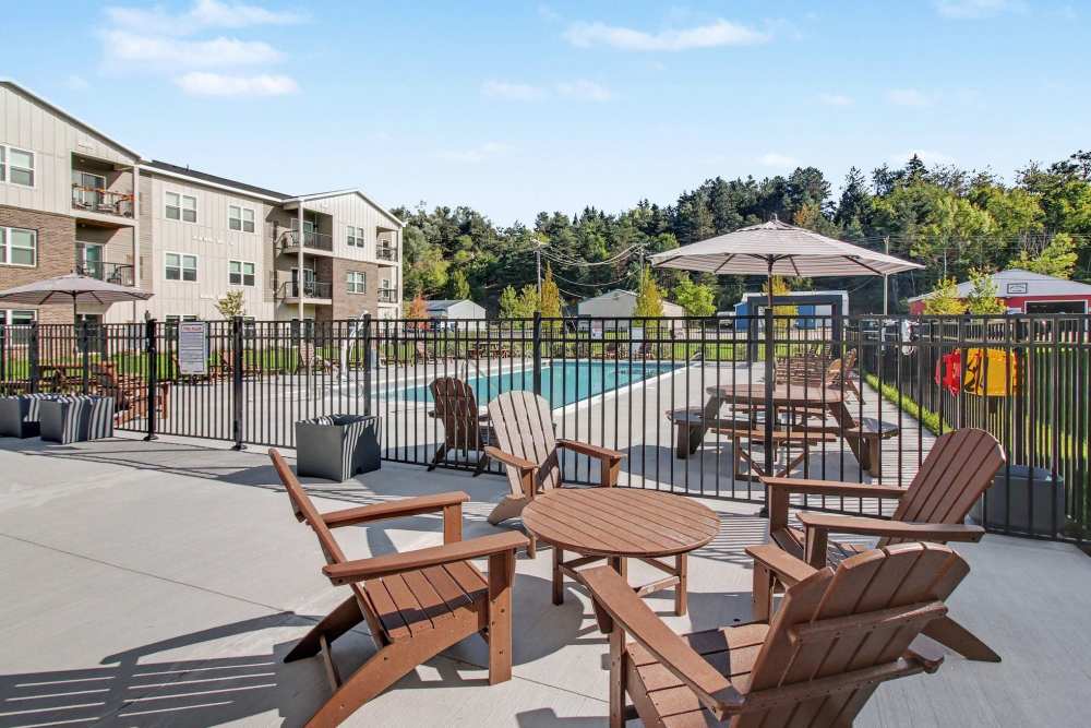 Poolside patio with brown Adirondack chairs and a round table, striped umbrellas, and a picnic table, enclosed by a black metal fence with apartment buildings in the background at The Block at Petoskey in Petoskey, Michigan