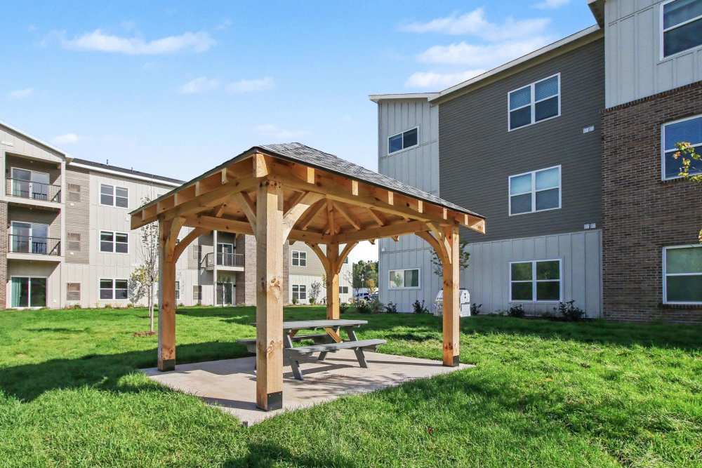 Wooden timber-frame gazebo with a shingled roof covering a picnic table, set on a grassy lawn near apartment buildings at The Block at Petoskey in Petoskey, Michigan