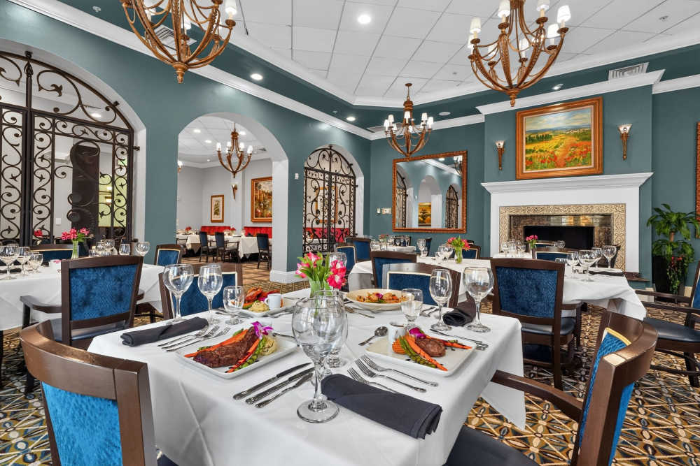 Elegant dining room with teal walls, set tables with plated entrées, chandeliers, wrought iron archways, and a fireplace at The Barclay at Vero Beach in Vero Beach, Florida