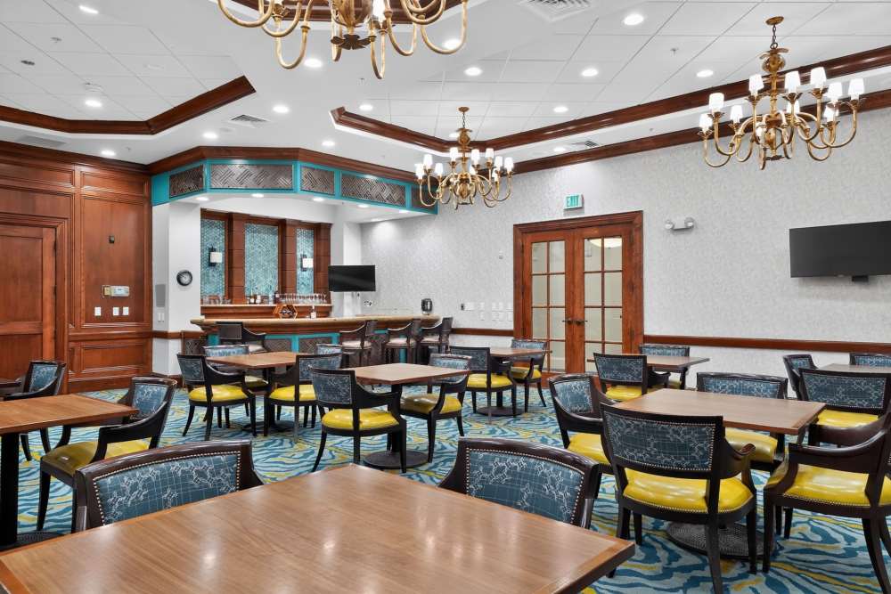 Social lounge with wood-topped tables, blue and yellow chairs, a bar area, and gold chandeliers at The Barclay at Vero Beach in Vero Beach, Florida