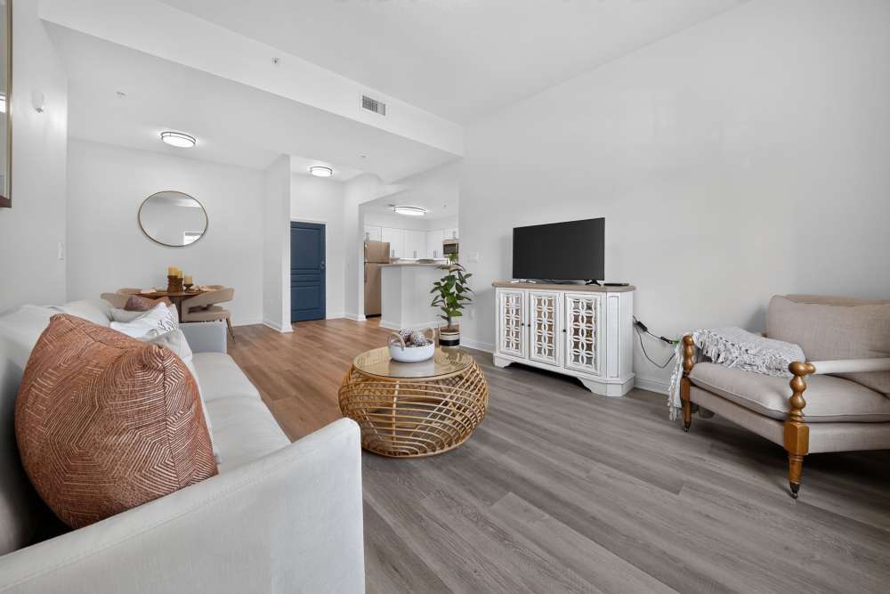 Bright apartment living room with a white sofa, rattan coffee table, TV console, and a view of the kitchen and dining nook at The Barclay at Vero Beach in Vero Beach, Florida