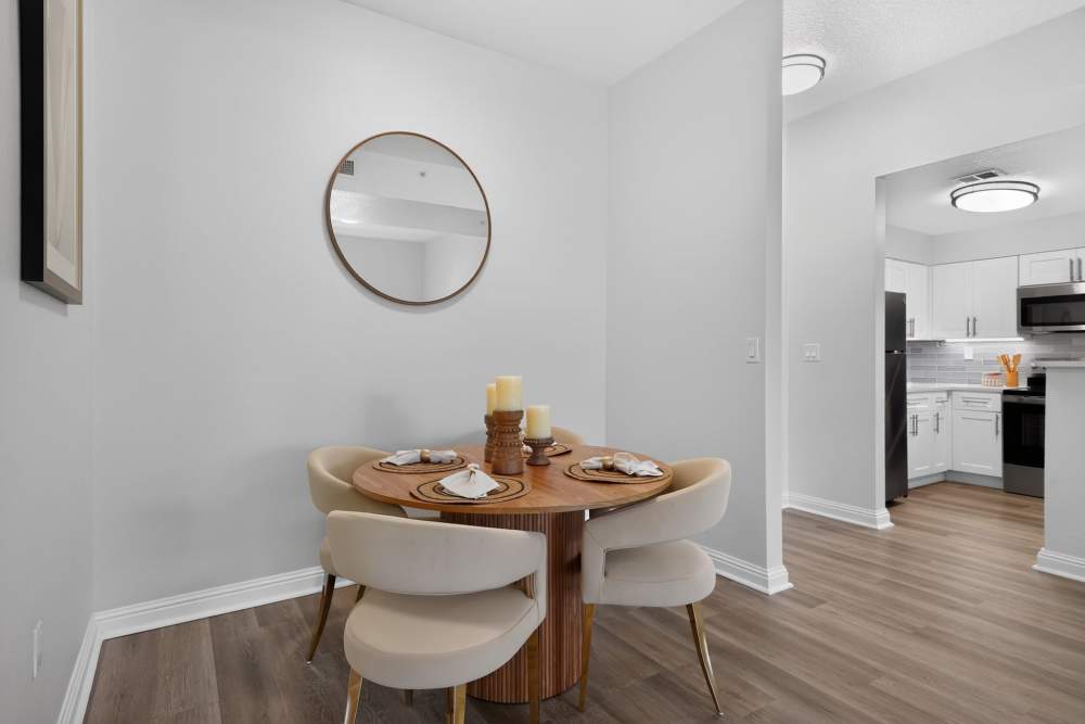 Apartment dining nook with a round wood table, four cream chairs, a round wall mirror, and a white kitchen beyond at The Barclay at Vero Beach in Vero Beach, Florida