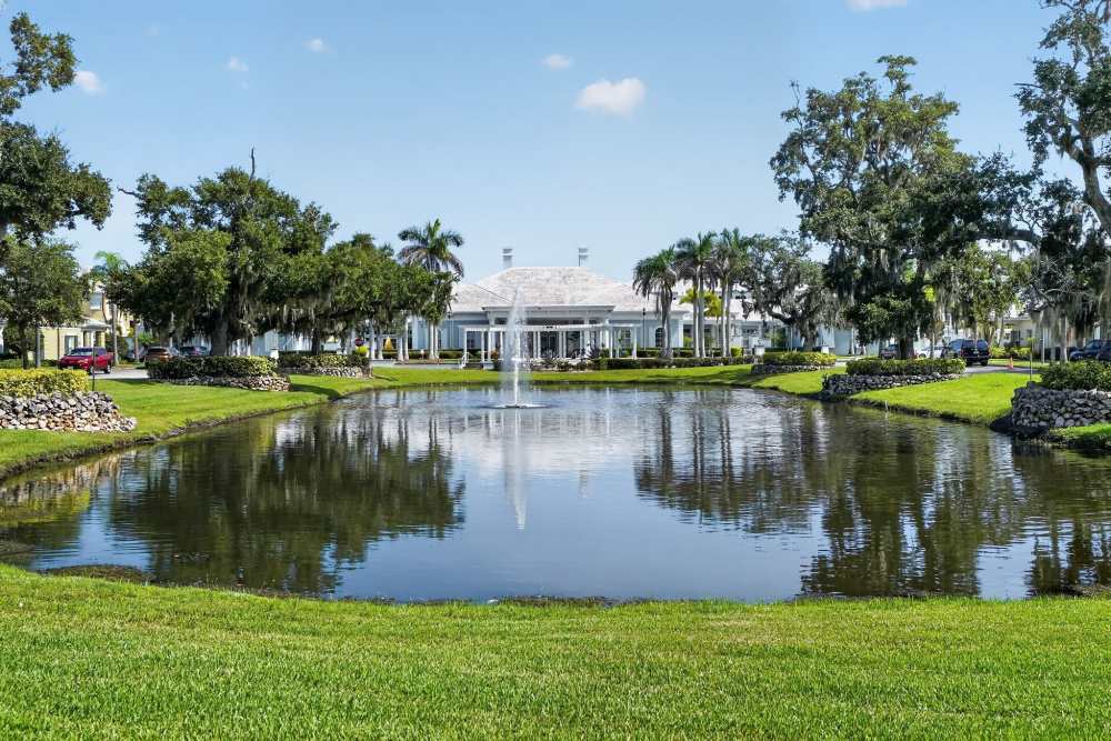 Community entrance with a fountain pond in the foreground, mature oak trees, and a white clubhouse building behind at The Barclay at Vero Beach in Vero Beach, Florida