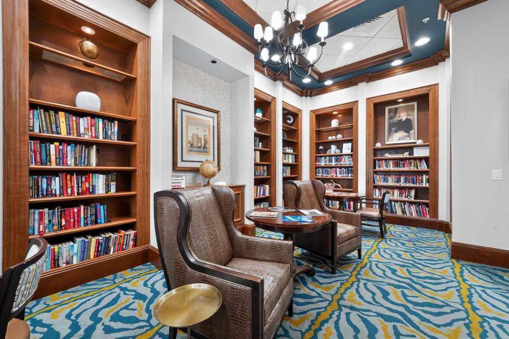 Library with built-in wood bookshelves, leather wingback chairs, a round reading table, and a patterned blue and yellow carpet at The Barclay at Vero Beach in Vero Beach, Florida