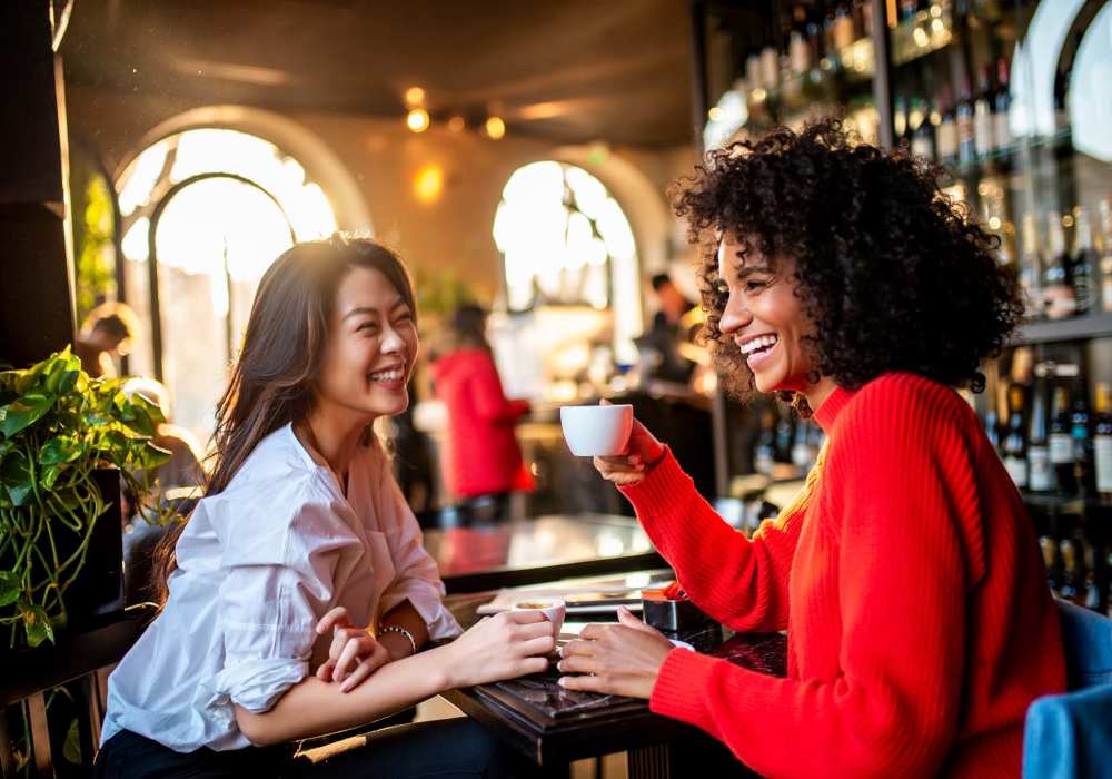 Two women enjoying coffee at a stylish cafe with plants and a warm atmosphere at Las Palmas Apartments in Brownsville, Texas