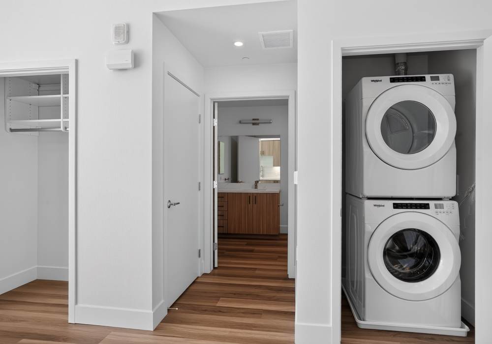 Stacked washer and dryer in closet beside shelving and hall to bathroom at The Colton in Redwood City, California