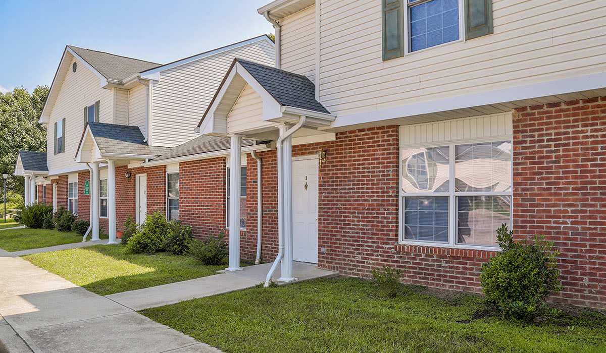 Row of apartment entrances with brick and siding exteriors, manicured lawns, and clear blue skies at Arlington Greene in London, Kentucky