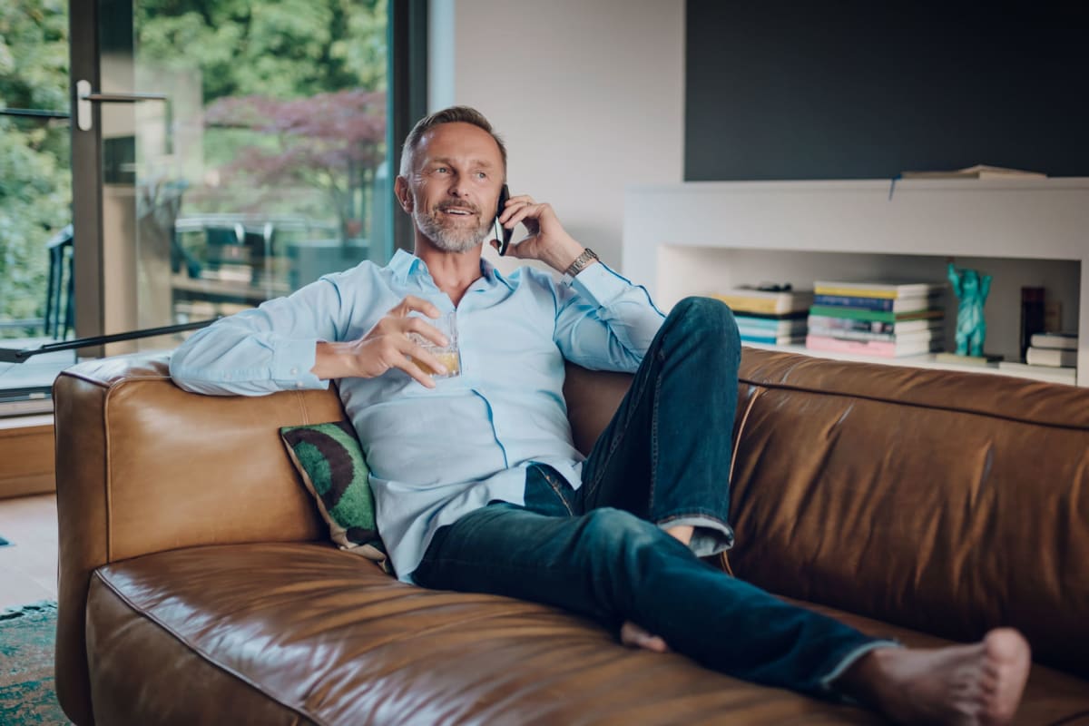 Resident relaxing with a drink on the couch while chatting with a friend on a mobile phone in his apartment at The Pointe at Siena Ridge in Davenport, Florida
