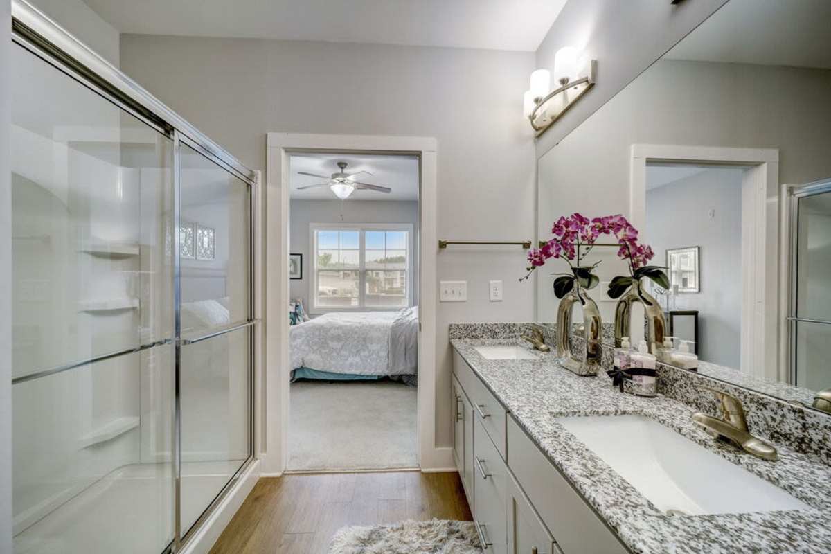 A model bathroom from a befroom showcasing the sinks and huge mirror with a flower decoration and a glass wall shower area at Century Park Place in Morrisville, North Carolina