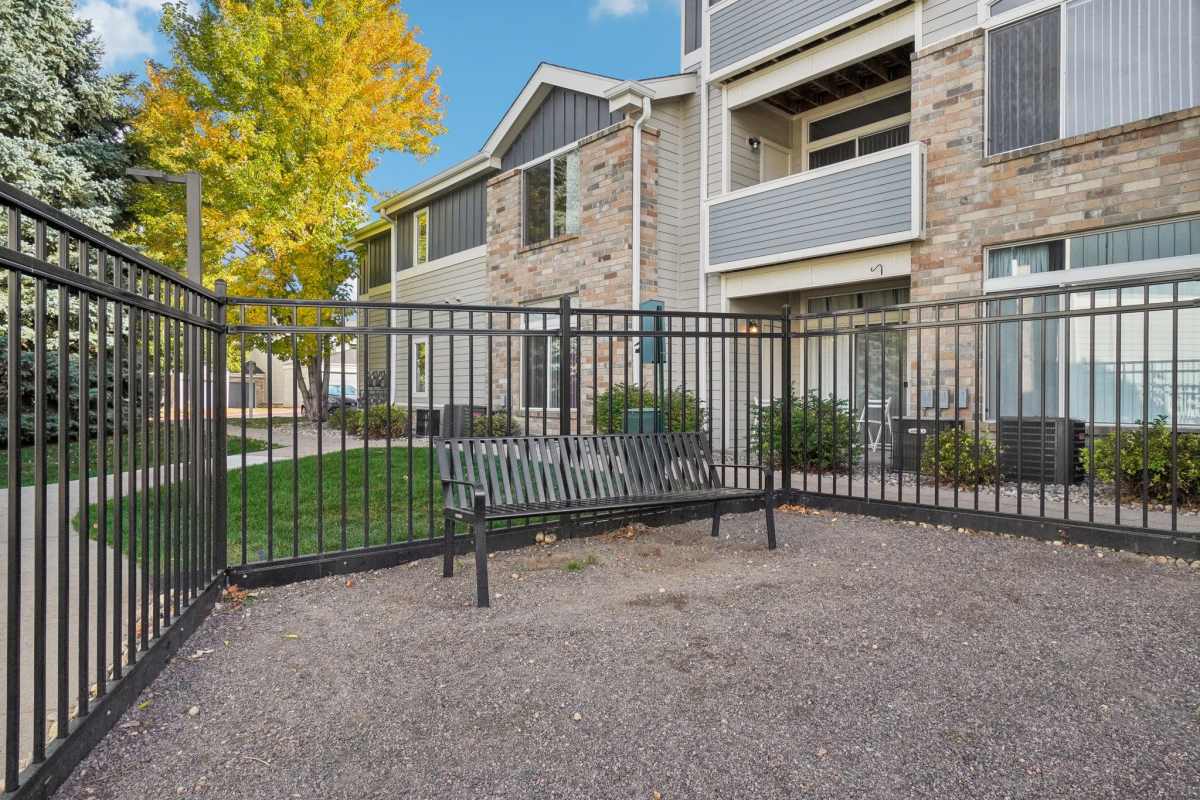 Fenced outdoor seating area with a wooden bench, gravel ground, and a view of a multi-story residential building with balconies at Pavilions at Silver Sage in Fort Collins, Colorado.
