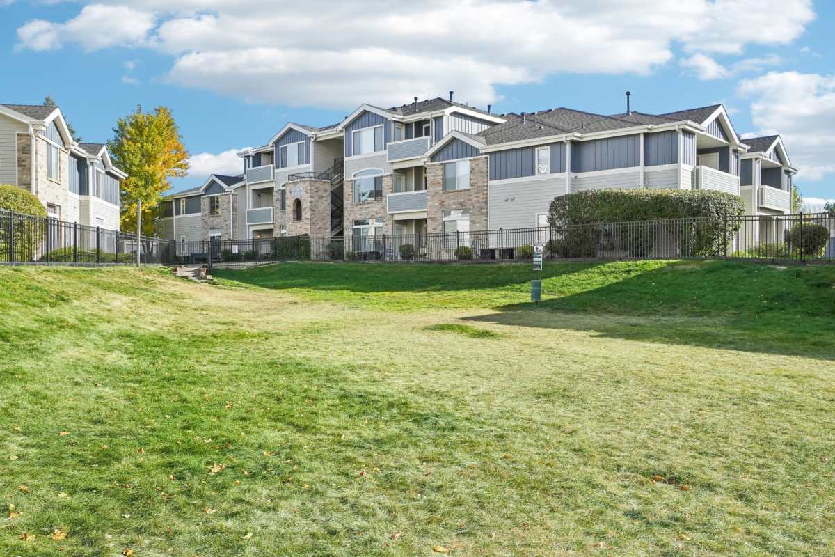 Modern apartment buildings with large windows, landscaped green space, and black metal fencing under a partly cloudy sky at Pavilions at Silver Sage in Fort Collins, Colorado.