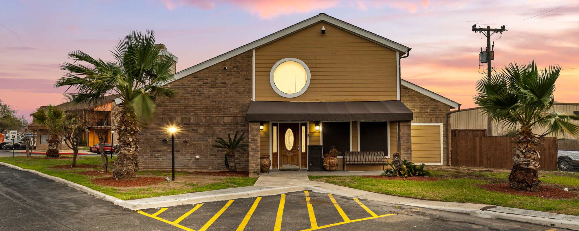 Gated entrance with palm trees and landscaped lawn at sunset at Towne East Village Apartment Homes in Converse, Texas